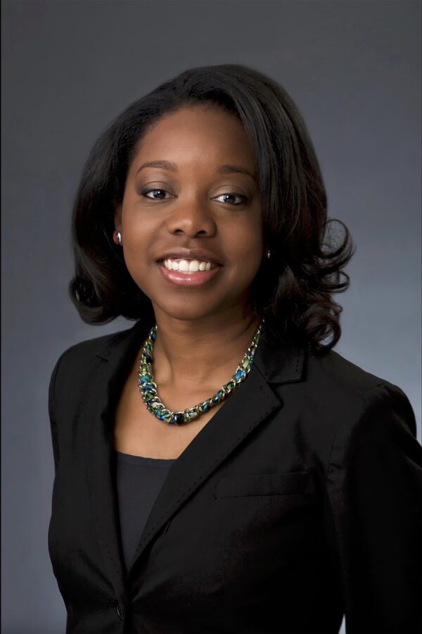 Young Black woman, lovely smile, with conservative black suit jacket over black sweater against a medium gray background.