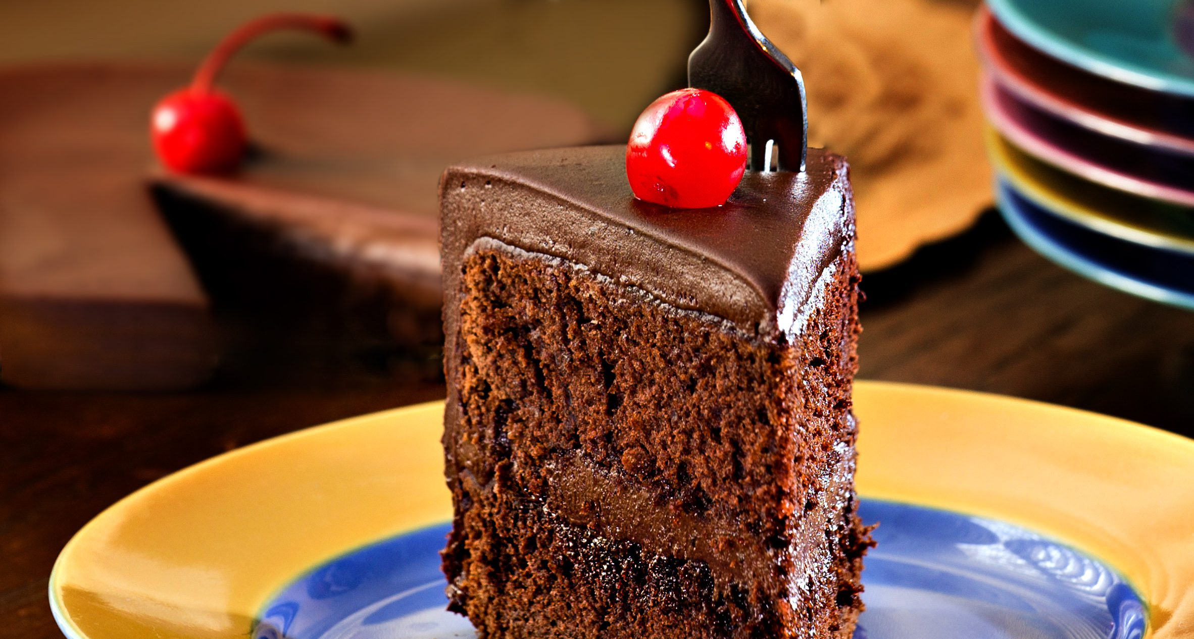 Rich chocolate cake, a maraschino cherry, a fork and colorful plates